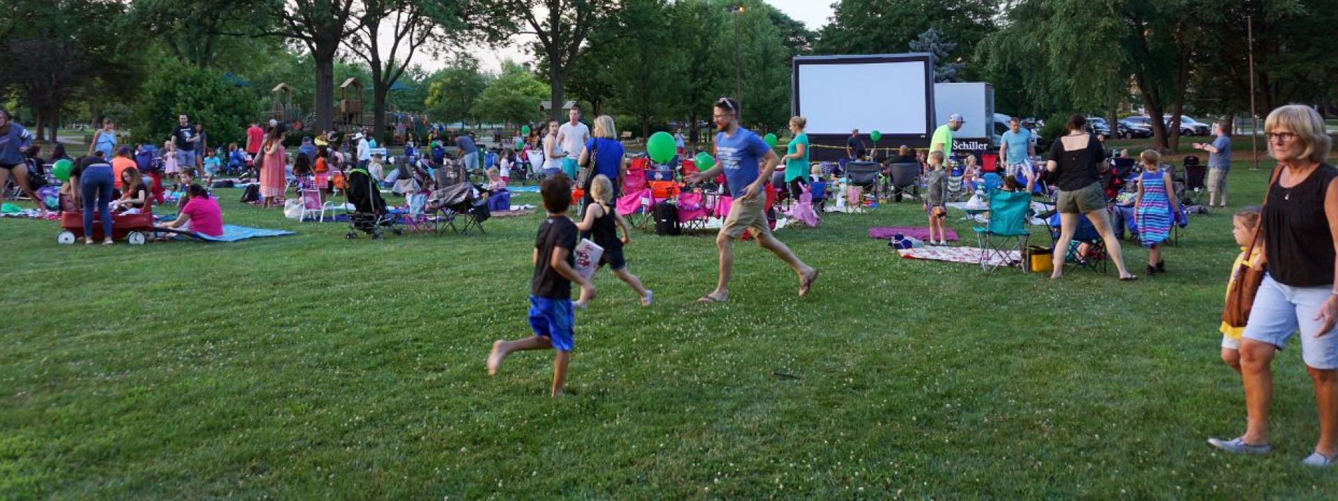 A large group of people are gathered on a grassy lawn watching an outdoor movie screen with trees in the background.