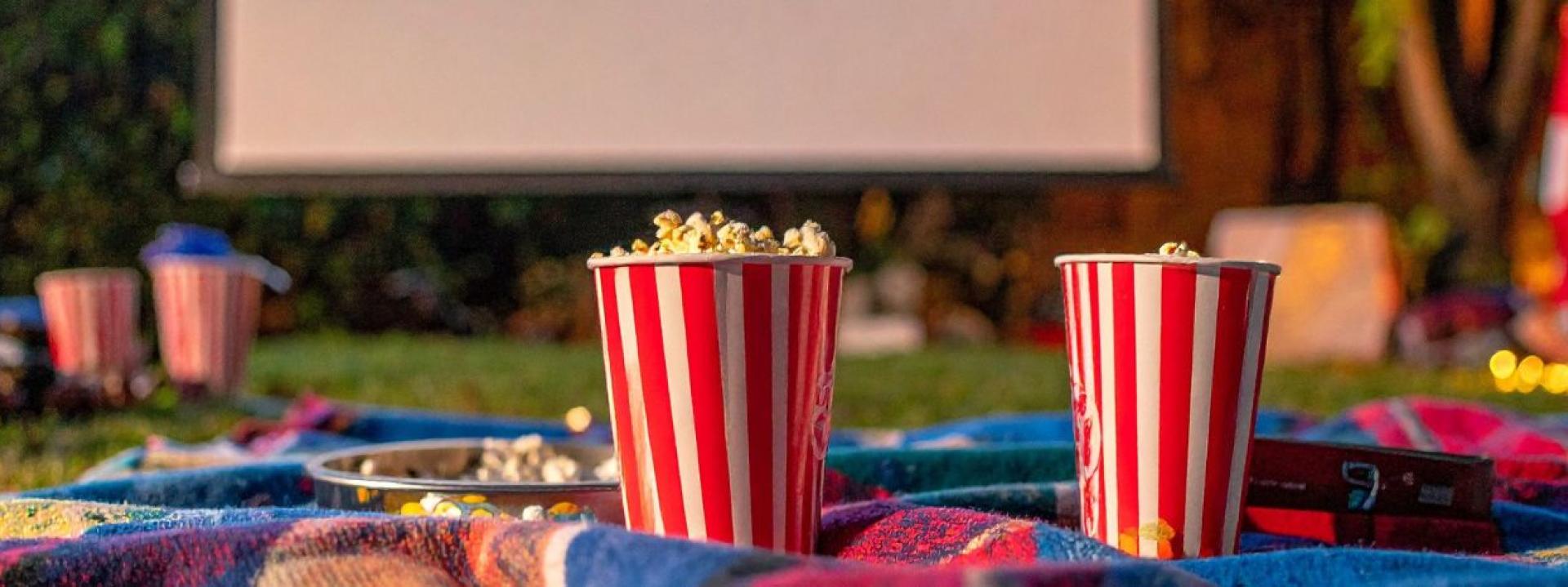 Two red-and-white striped popcorn buckets on a colorful outdoor picnic blanket with scattered snacks in the foreground and a movie screen in the background.