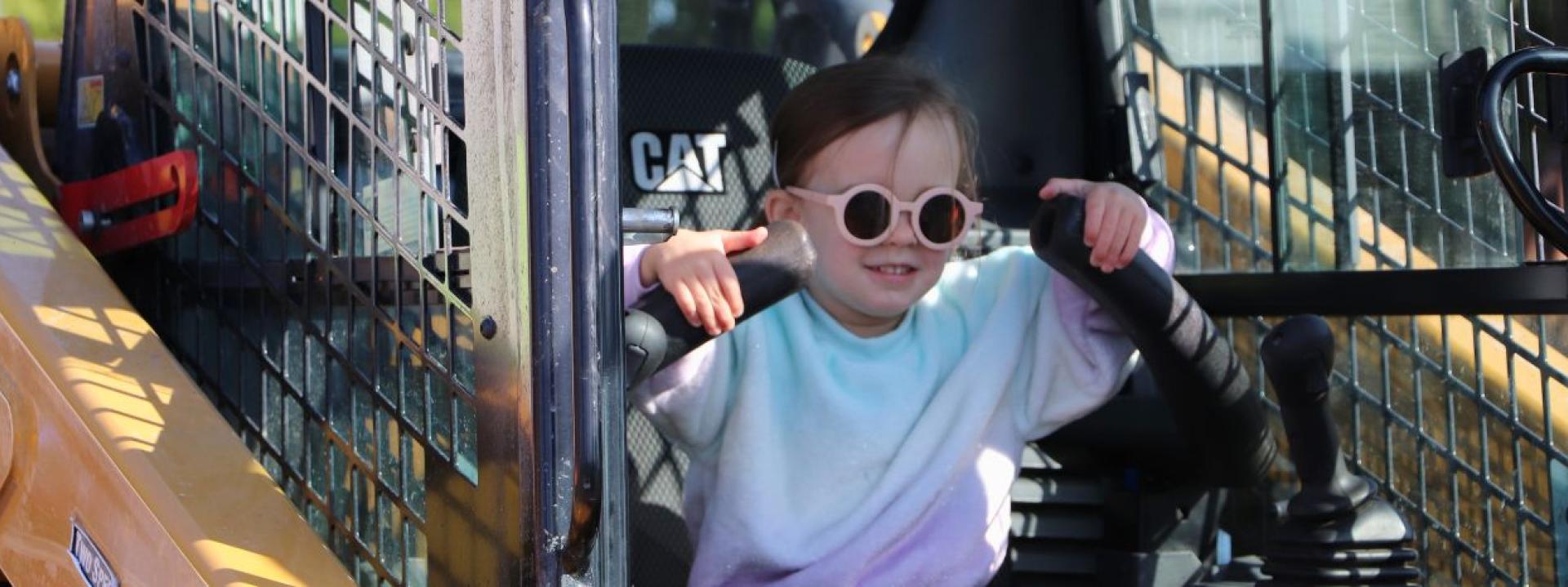 A little girl with sunglasses smiles which sitting in a skid steer