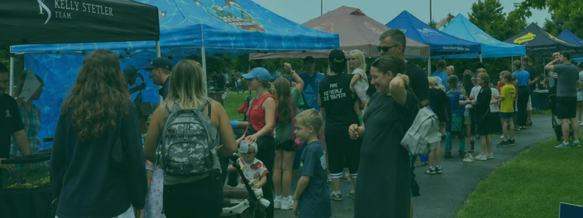 Outdoor community event with pop-up tents and a crowd gathered along a walkway, with families and children standing near the booths.