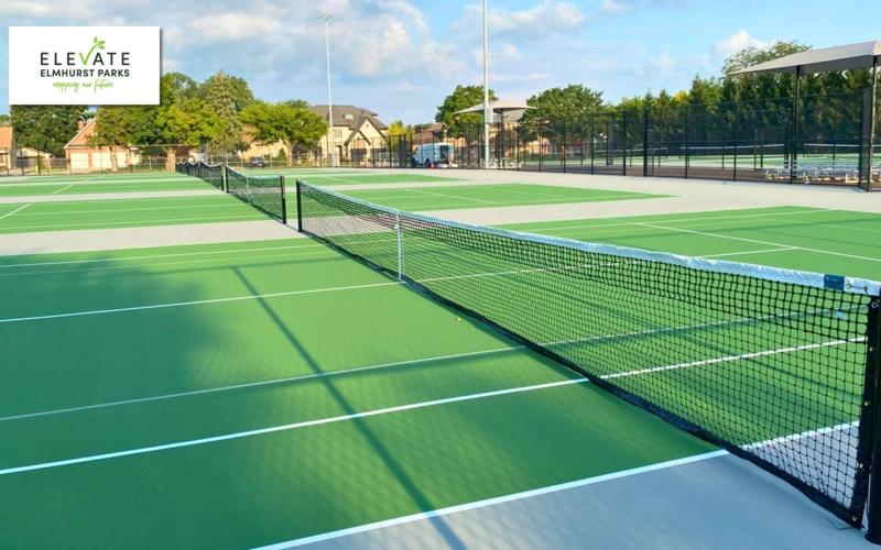 Outdoor tennis court with green hard surface, white lines, and a black tennis net. Shade structures and bleachers are in the background.