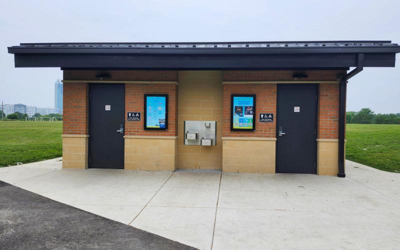 A brick restroom building with two dark doors, a water fountain, set against a grassy field and sky.