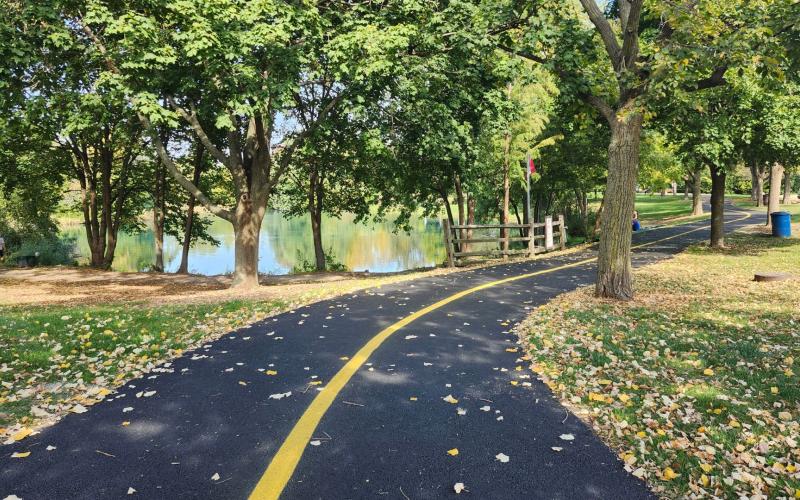 A curved blacktop path with a yellow center line runs through a park lined with leafy green trees. A body of water is seen in the background.