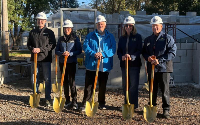 Five people wearing hard hats pose outdoors holding shovels in front of a construction site with gravel ground.