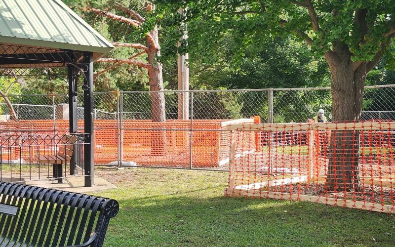 Orange construction fencing in a park. To the left is a green roofed gazebo. 