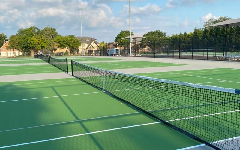 A group of tennis courts with fencing behind. Beyond the fencing is a shade structure.