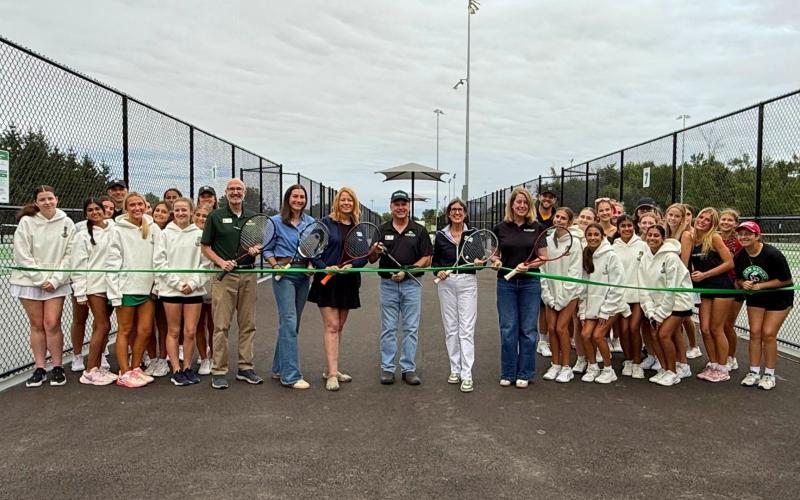 A group of people stand between tennis courts. They hold a ribbon.