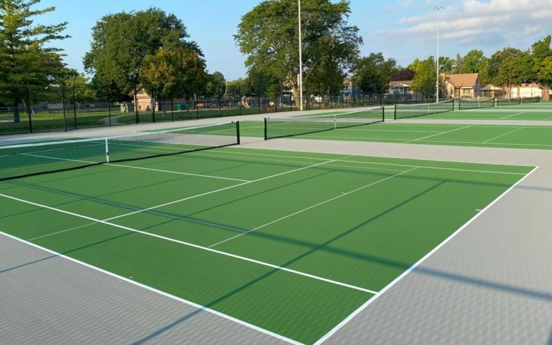 Outdoor tennis court with green playing surface, white boundary lines, and chain-link fencing, viewed from the sideline.