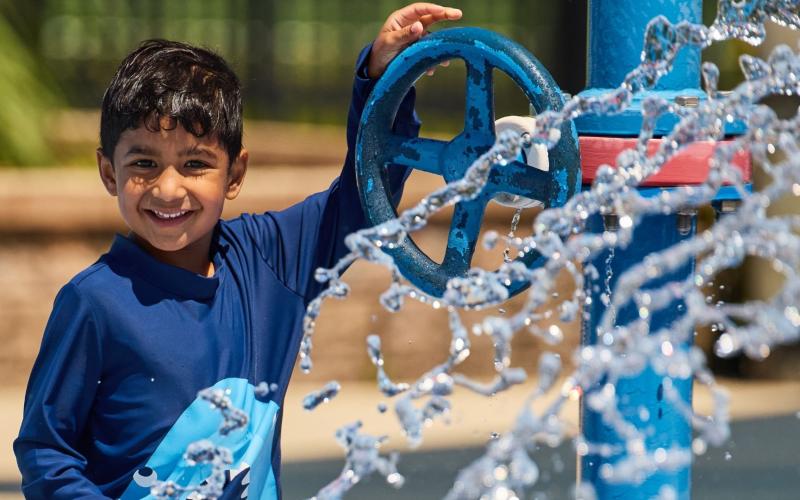 A smiling child in a blue swim shirt stands beside a blue water sprinkler, with water splashing and sparkling in the foreground.