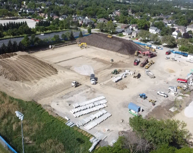 Aerial view of a construction site with heavy machinery, piles of dirt, and stacks of pipes. Buildings and trees are visible in the background.