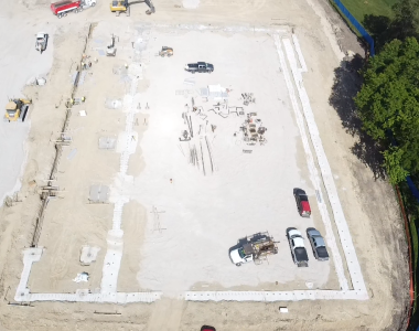 Aerial view of a construction site with heavy machinery, vehicles, and building materials on a dirt and gravel foundation next to a wooded area.