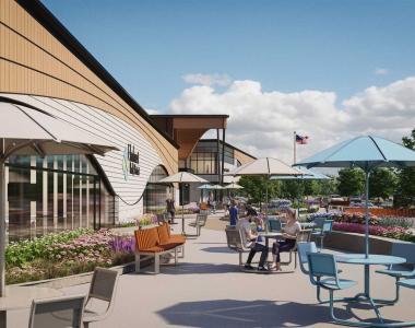 Outdoor modern rendering of a large building with curved glass facade, landscaped planters and benches, several people walking and sitting, and cars visible in a parking lot.