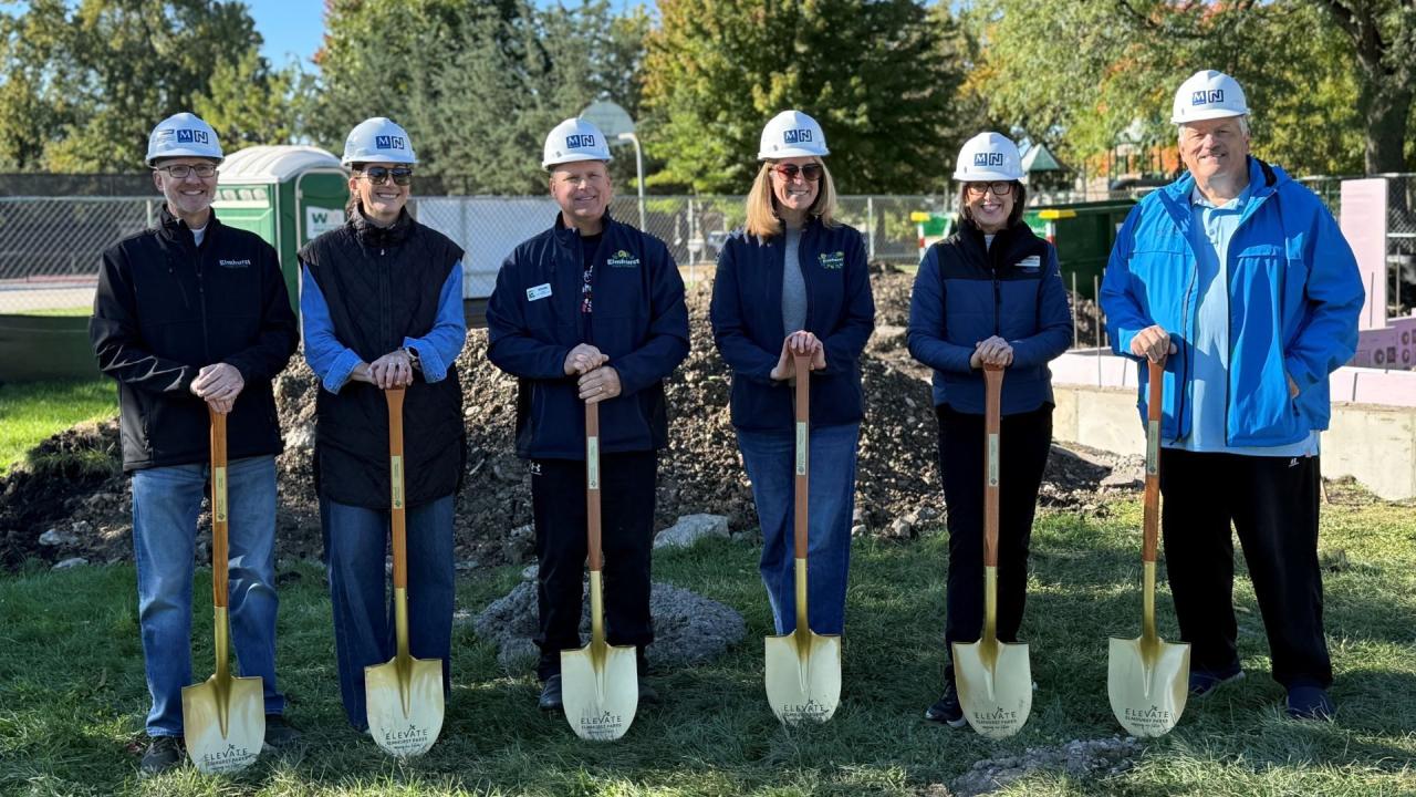 Seven people in hard hats stand outdoors in a row holding shovels planted in the ground, with a green lawn, trees, and fencing in the background.