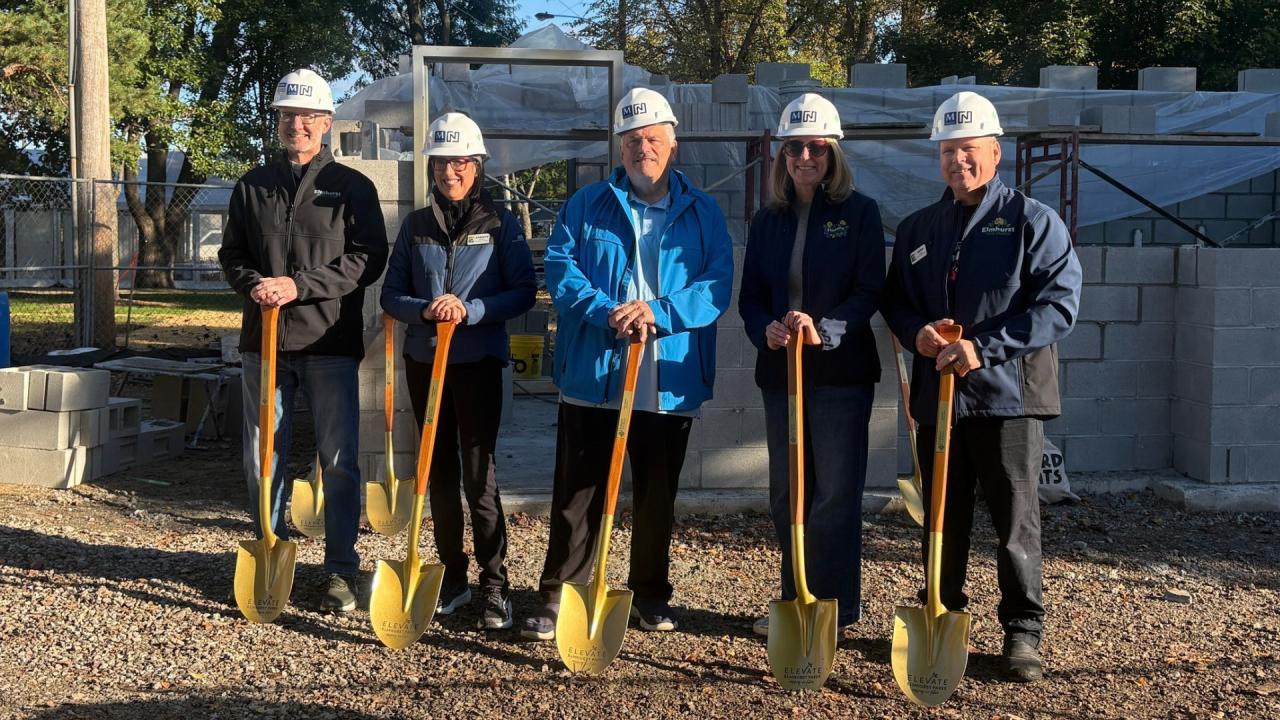 Five people wearing hard hats pose outdoors holding shovels in front of a construction site with gravel ground.
