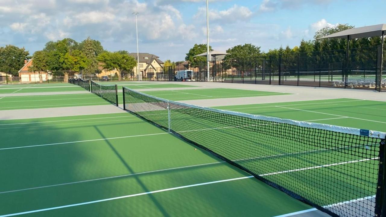 Green outdoor tennis courts with white lines, black nets in the foreground, and shade structures in the background.