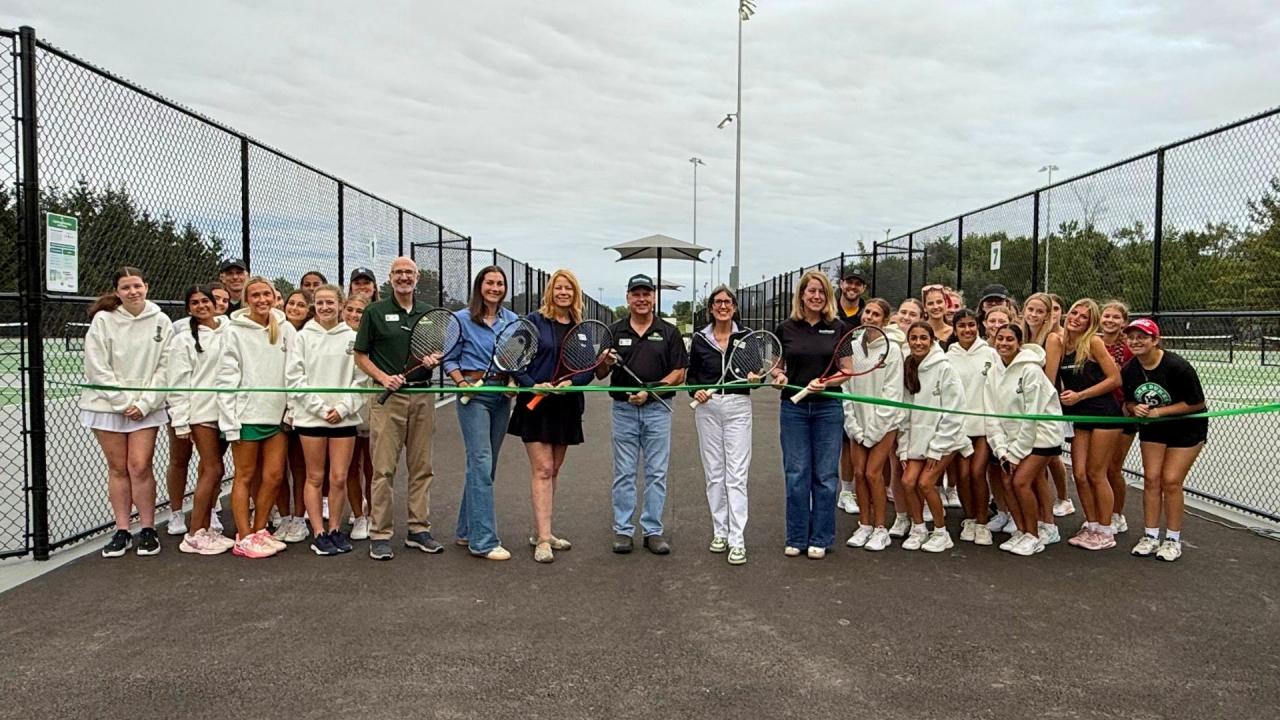 A group of people stand between tennis courts. They hold a ribbon.