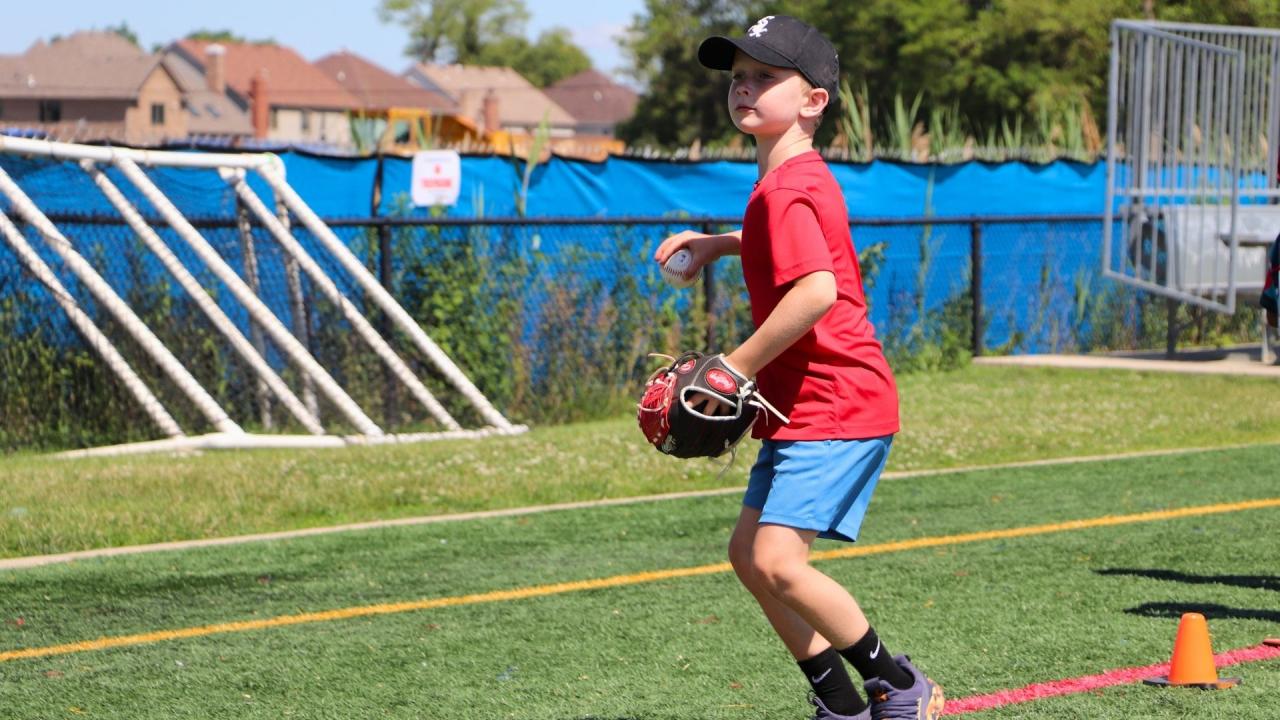 A child stands holding a baseball on a green outdoor turf field, wearing a baseball cap and holding a small glove, getting ready to throw.