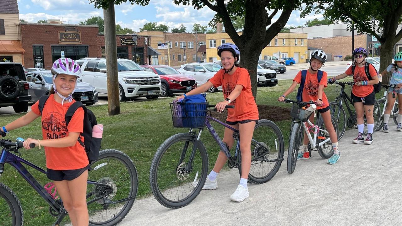 A group of kids wearing helmets and orange shirts stand with bicycles on a trail, with cars and buildings in the background.