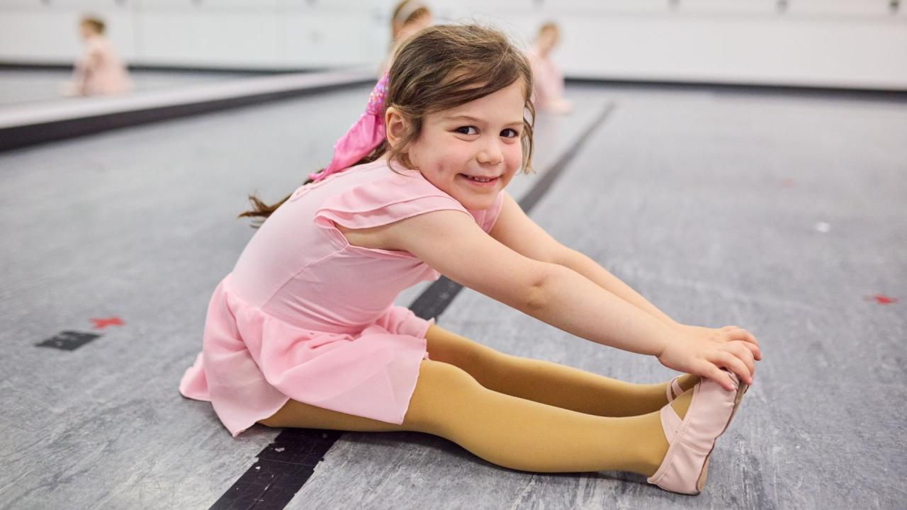 A child in a pink ballet outfit on a dance floor doing a stretching exercise, smiling and reaching forward to touch her toes.