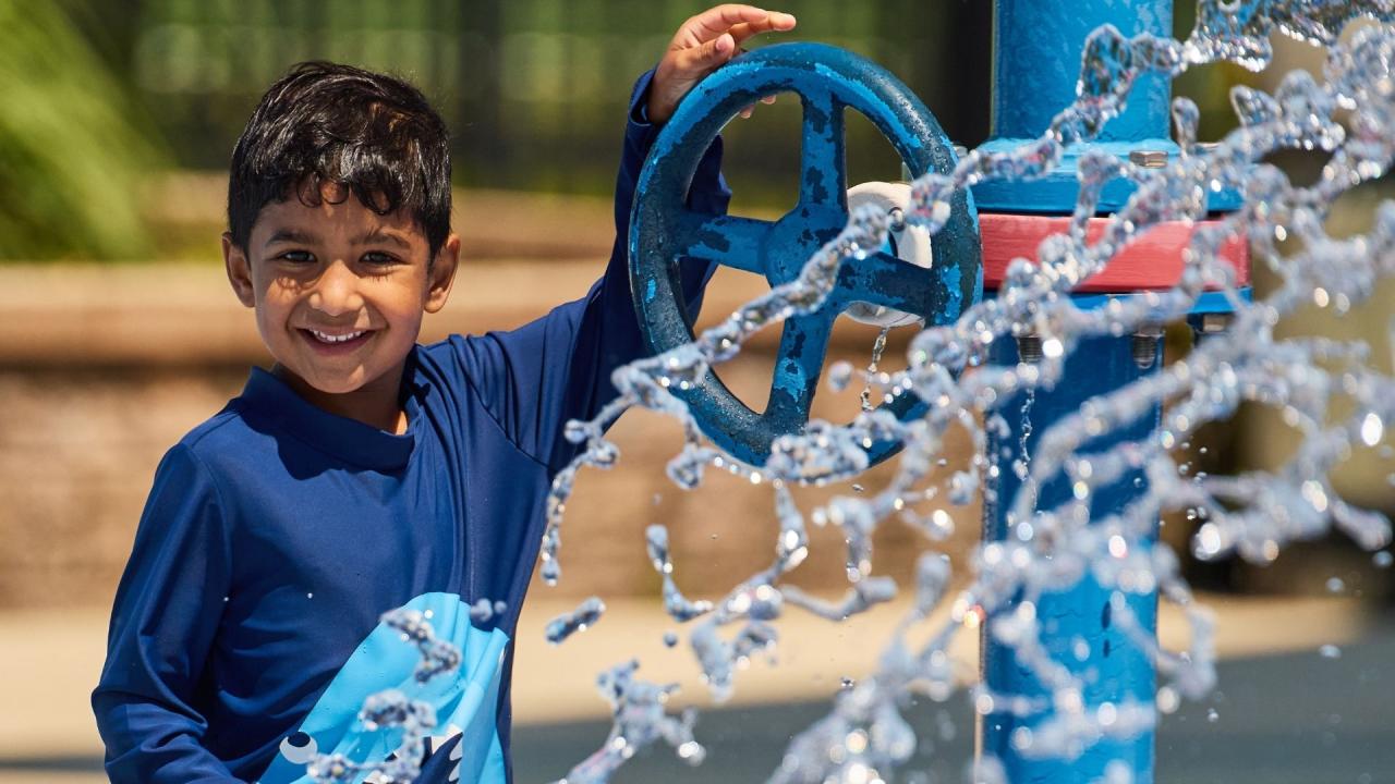 A smiling child in a blue swim shirt stands beside a blue water sprinkler, with water splashing and sparkling in the foreground.