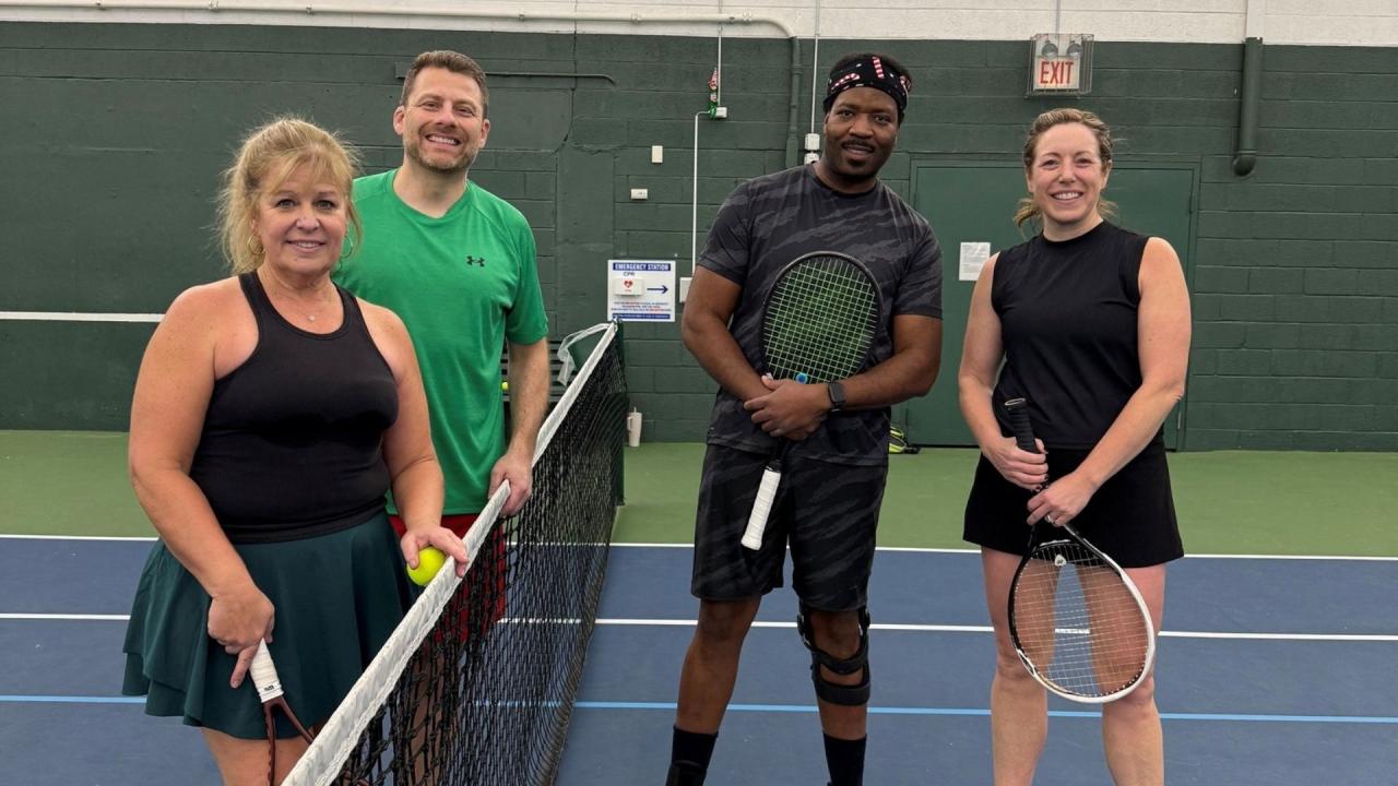 Four people on an indoor tennis court posing with rackets, smiling toward the camera
