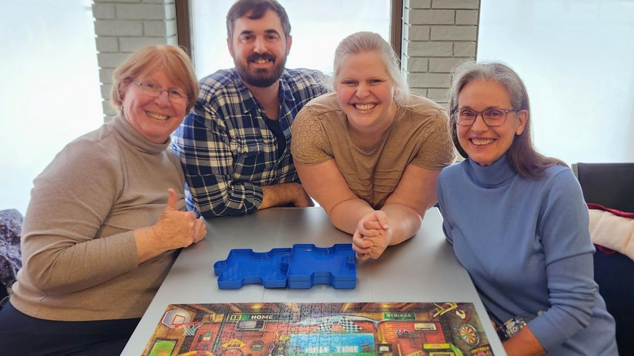 Four people smiling at the camera around a table with a puzzle and a blue puzzle sorter.