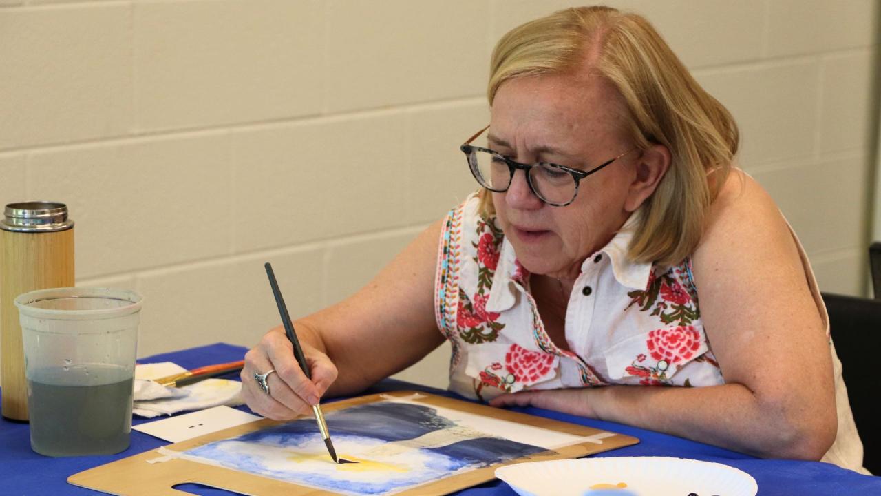 A person wearing glasses and a floral-patterned shirt is painting on a watercolor pad with a brush and water cup on the table.