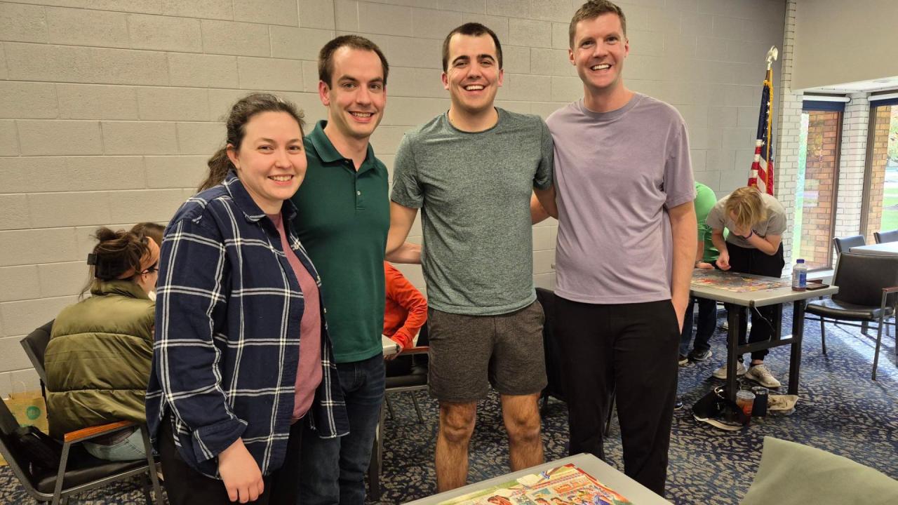 Four people stand and smile while others work on puzzles in the background.