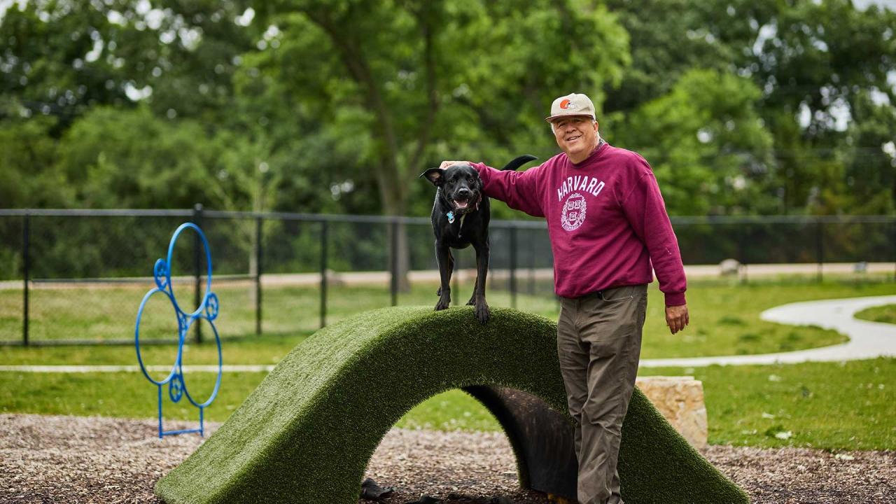 A person in a maroon sweatshirt and a baseball cap stands next to a black dog on an agility ramp in a park.