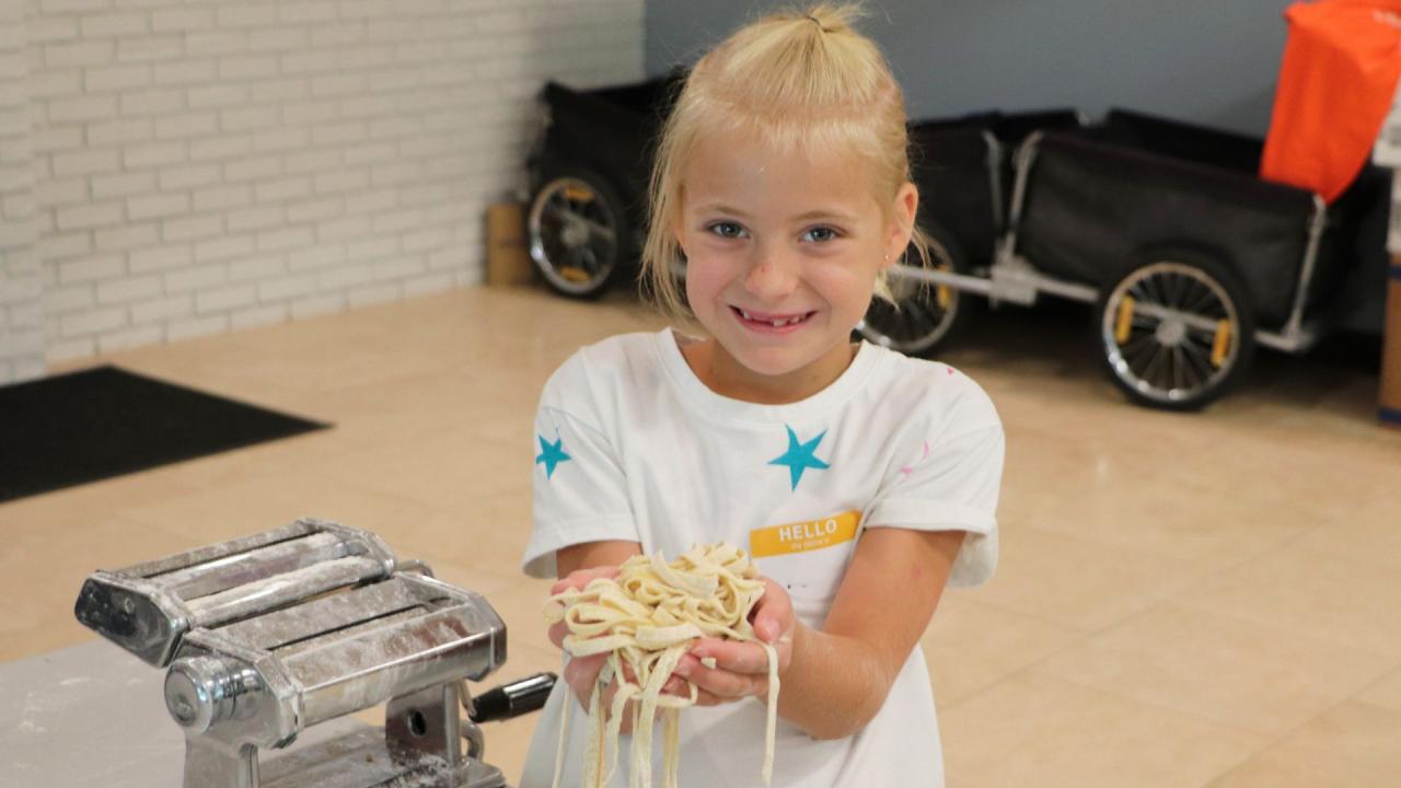 A young person smiles while holding a pile of freshly made pasta in their hands next to a pasta maker.