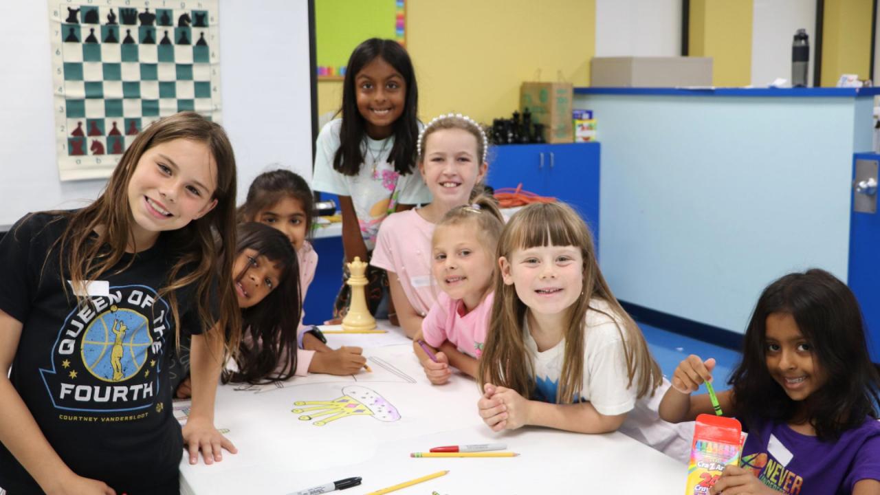 A group of children are gathered around a table with art supplies. A chessboard is visible on the wall behind them.