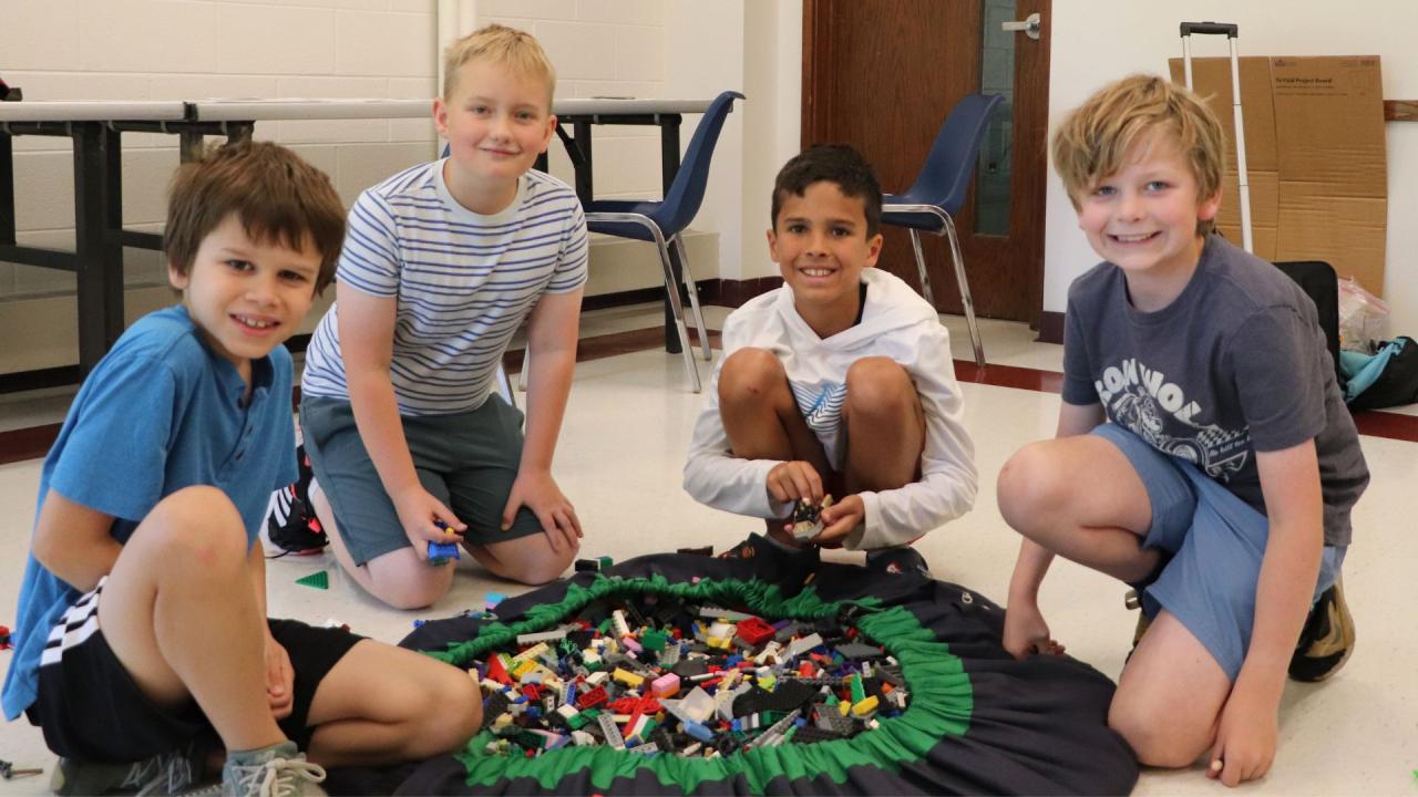 Four children are crouching around a large circular mat filled with LEGO bricks, smiling at the camera.
