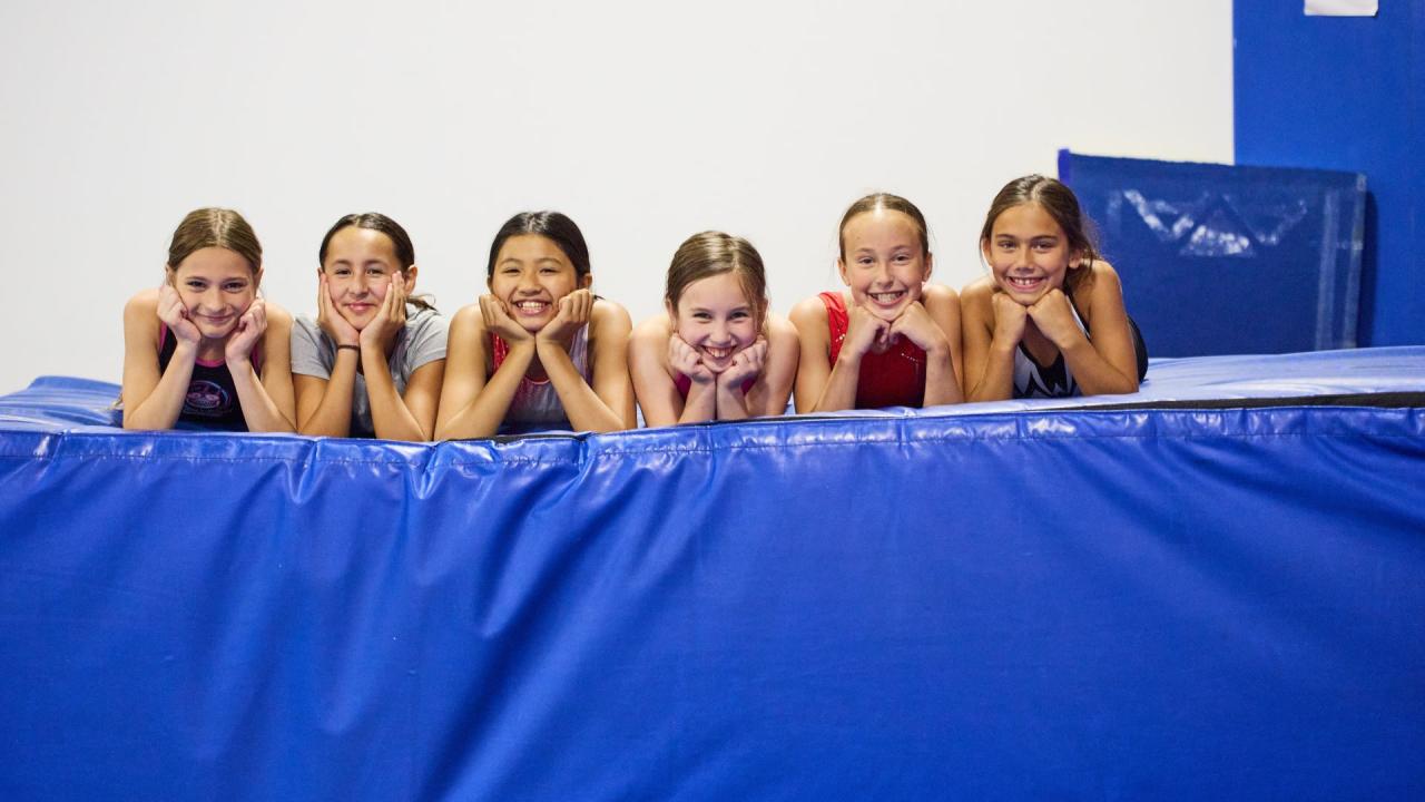 Six smiling children lying on a blue mat in a gymnastics facility.