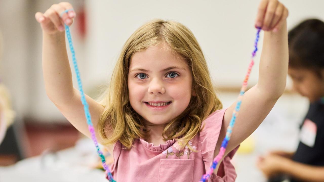 A child smiles and holds up a colorful beaded necklace they have made.