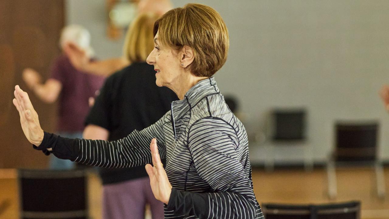 A woman practices Tai Chi with her hands raised in front of her. Other people are visible in the blurred background.