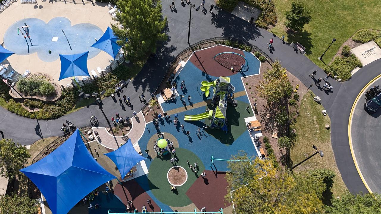 Aerial view of a vibrant playground with blue sunshades, green play structures, and a splash pad area with people enjoying the space.