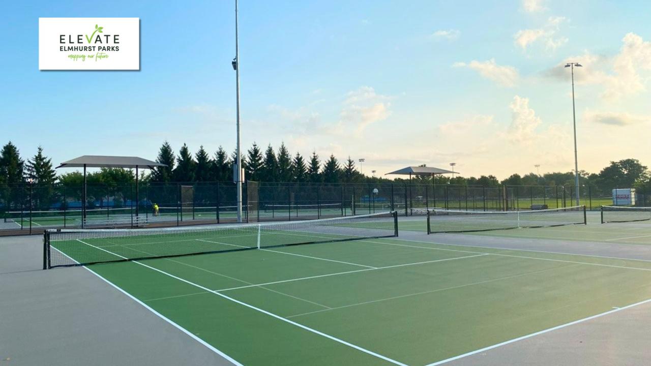 Refinished tennis courts Outdoor tennis court with green playing surface, white boundary lines, and chain-link fencing, viewed from the sideline with tall light poles and shade structures in the background.