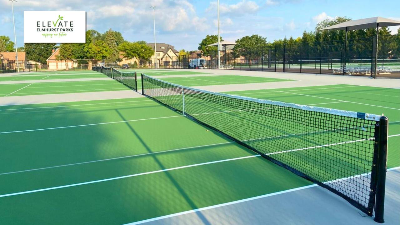 Refinished tennis courts Outdoor tennis court with green hard surface, white lines, and a black tennis net. Shade structures and bleachers are in the background.