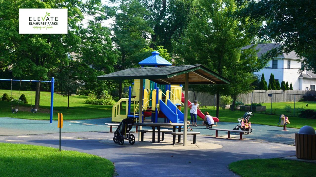 A playground with a colorful climbing structure, slides, swings, and picnic tables surrounded by green trees and grass.