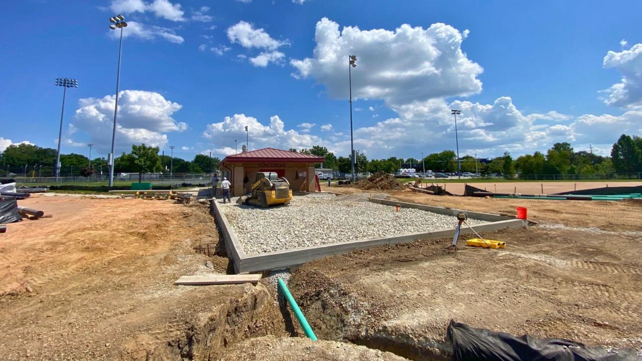 Prep work for new shade structure and paver patio, August 2026. Construction site with gravel and concrete footing in the foreground, tall light poles, a small red shed.