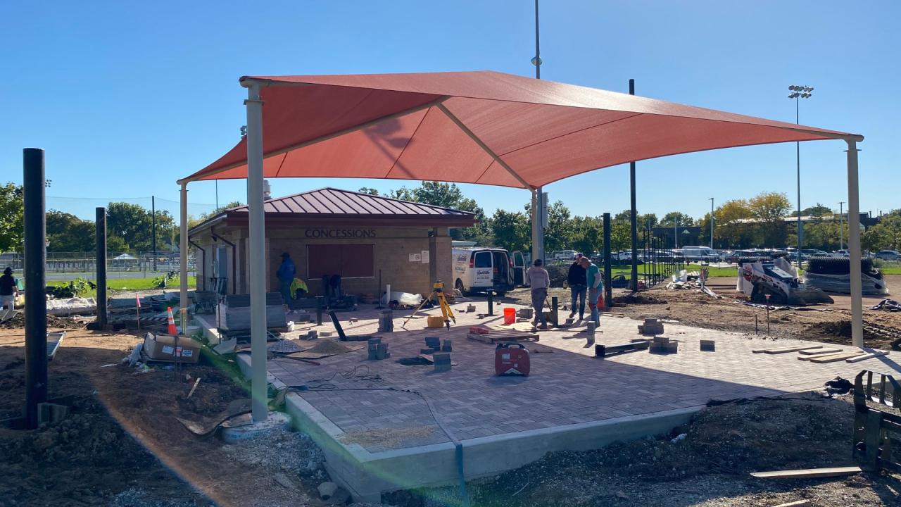Central shade construction, October 2026. Outdoor construction site with a built building, a large red canopy on metal posts, scattered tools and workers standing near equipment.
