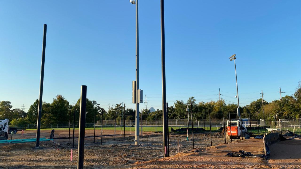 Installation of poles for the new netting backstops and fence, October 2026. Outdoor sports field under construction with dirt ground, scattered equipment and poles, small lights on tall standards, and a clear blue sky.