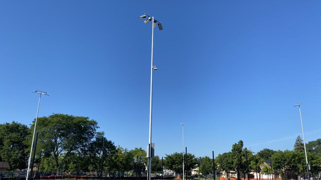 New light poles at the South Tennis Courts Tall light poles stand against a clear blue sky, with trees and buildings in the background.
