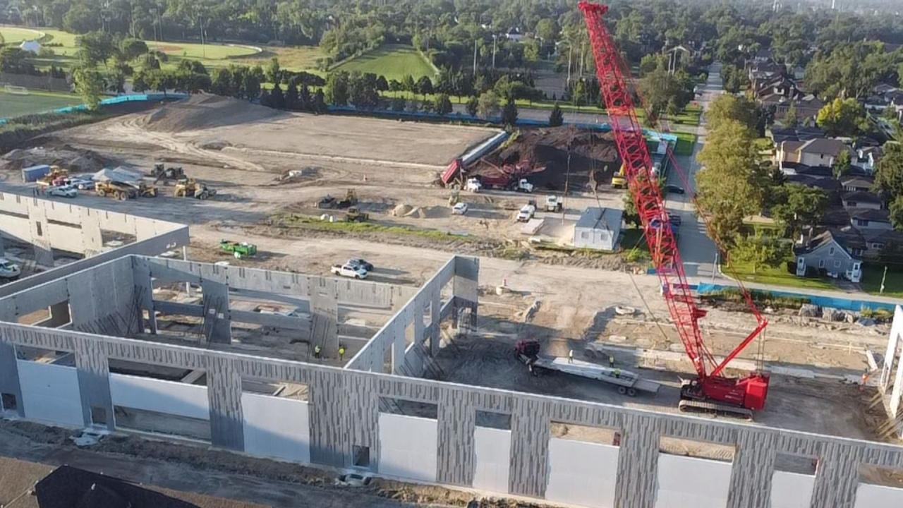 Aerial view of a construction site with partially built concrete building frames and a tall red crane, surrounded by trees, houses, and fields.