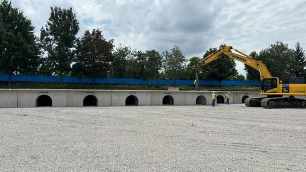 A yellow excavator is parked next to a a StormTrap structure at the Wagner Community Center site with multiple arched openings. Several people in hard hats are standing near the structure.